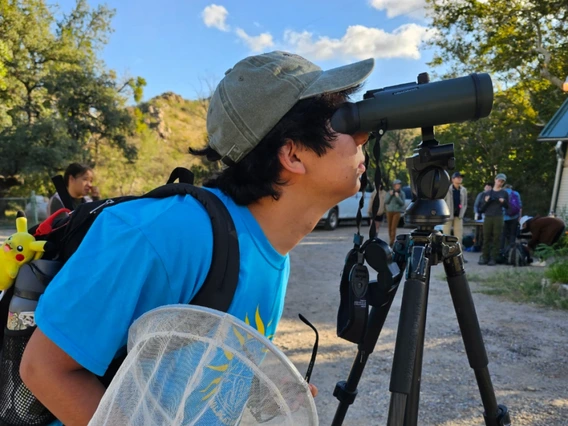 U of A student looks through binoculars and holds a insect net during the 2024 BioBlitz at the SRER.